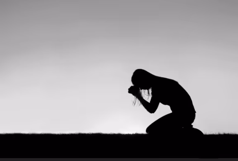 Silhouette of a woman kneeling in prayer against a bright sky