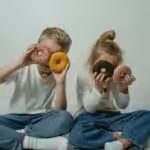 Two children sitting indoors, playfully holding colorful sprinkled doughnuts to their eyes.