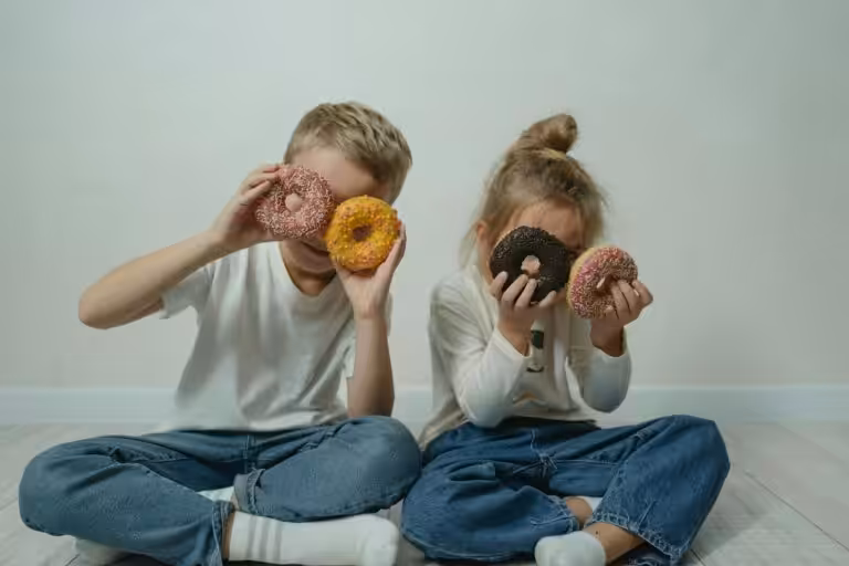 Two children sitting indoors, playfully holding colorful sprinkled doughnuts to their eyes.