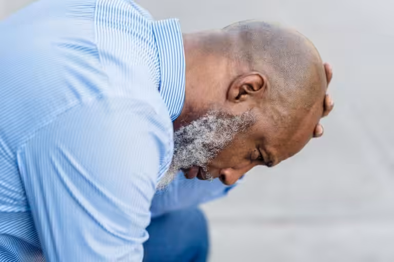 A middle-aged man in a suit looks distressed while sitting outside, hand on head.