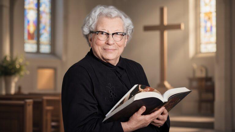 Elderly woman in a church holding an open Bible, smiling.